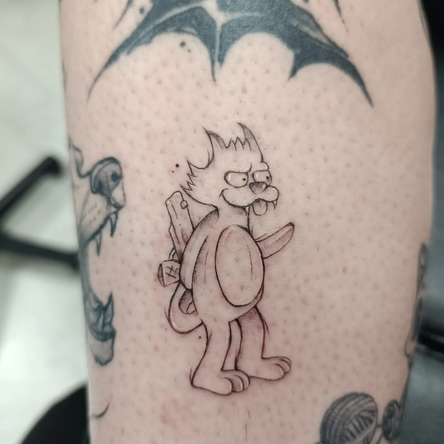 Tatouage fine line d’un petit personnage de dessin animé, encre noire sur bras tatoué, par Sophie Guiot